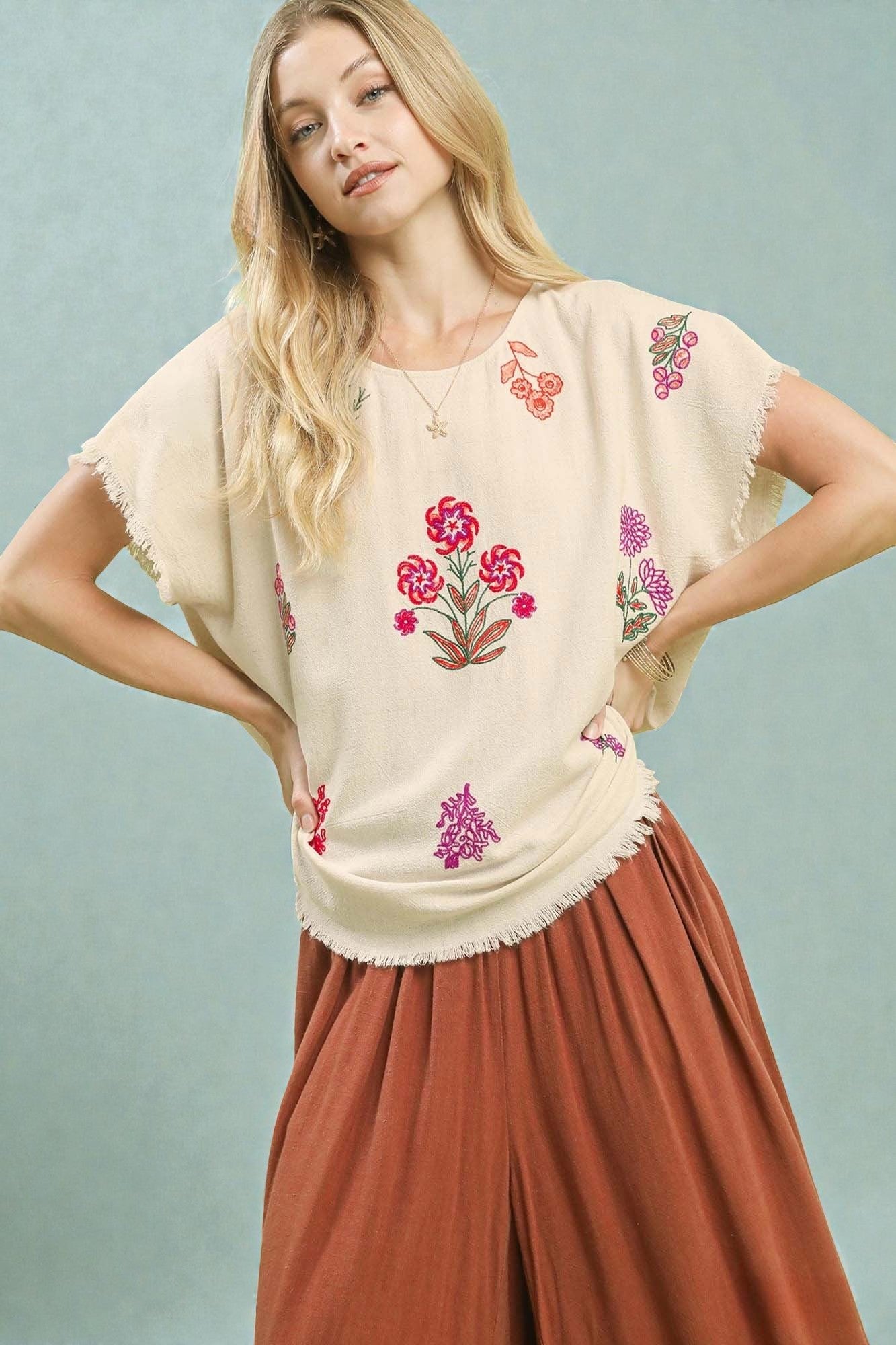 Woman wearing a beige top with floral embroidery and brown pants against a light blue background