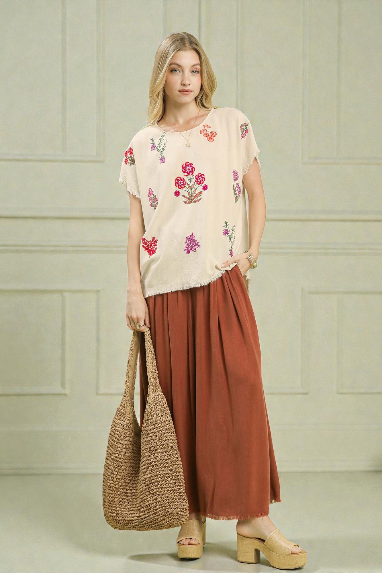 Woman wearing a floral top and rust-colored pants, holding a straw bag against a light green wall.