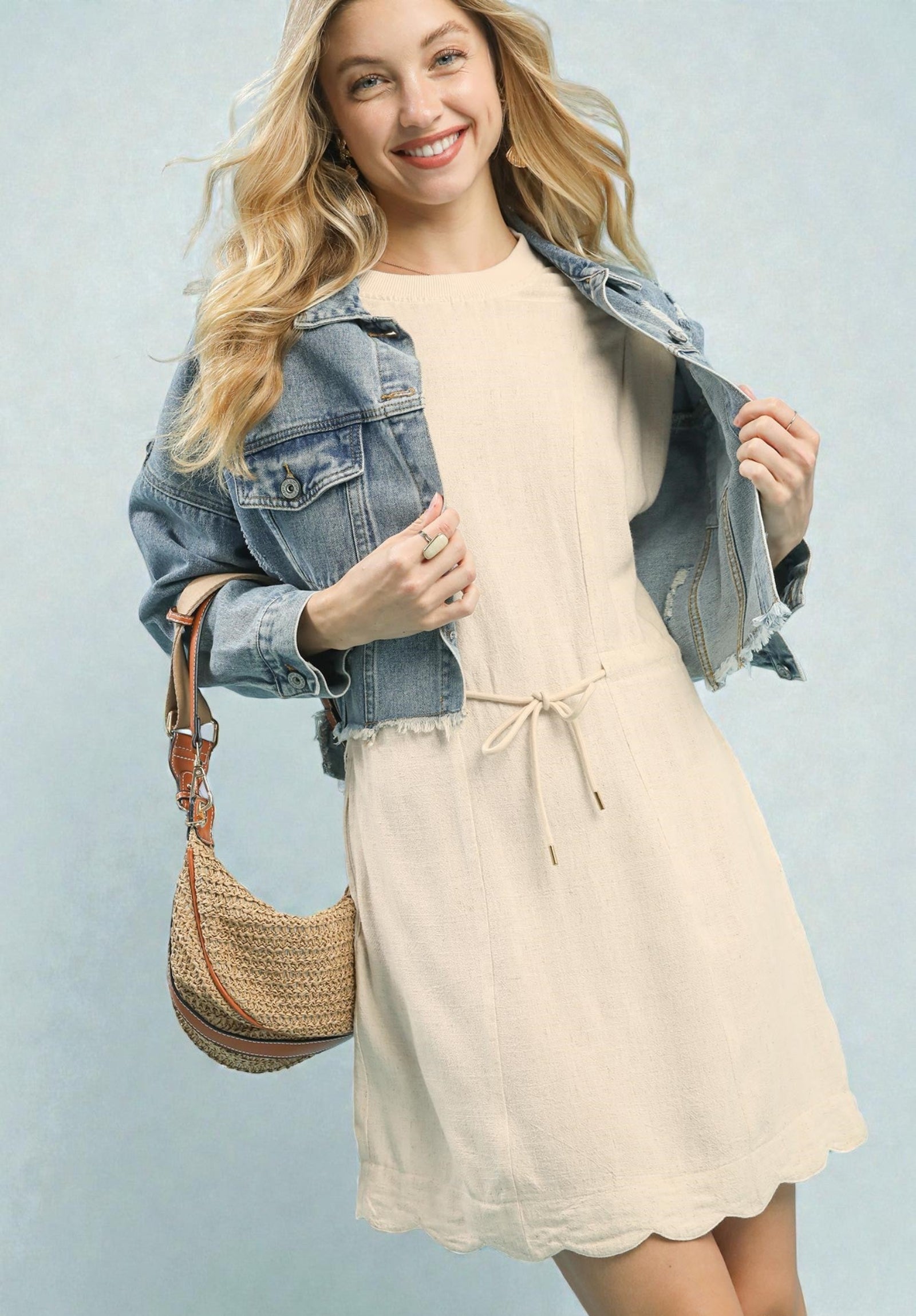 Woman wearing a beige dress with a denim jacket and holding a woven bag against a light blue background