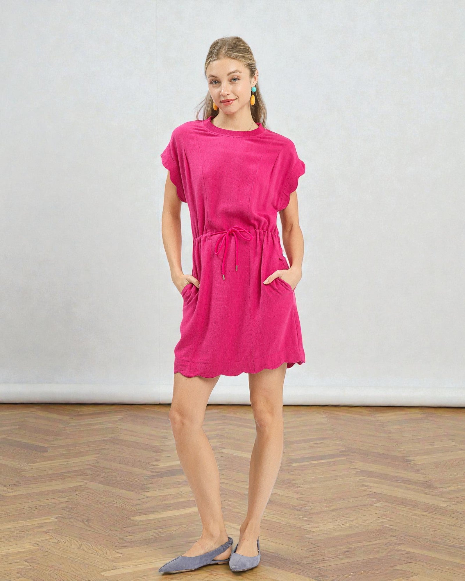 Woman wearing a bright pink dress standing on a wooden floor with a white wall background
