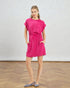 Woman wearing a bright pink dress standing on a wooden floor with a white wall background