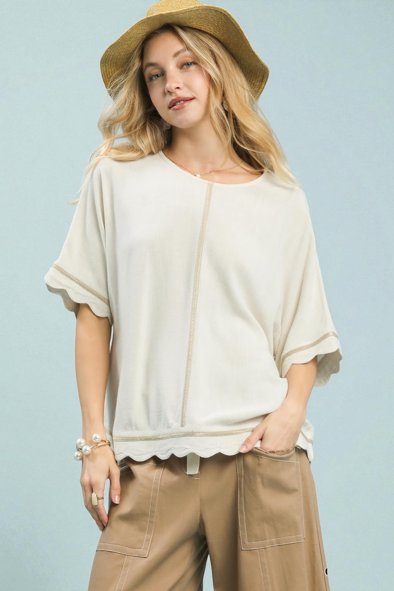 Woman wearing a beige hat, white top, and brown pants against a light blue background