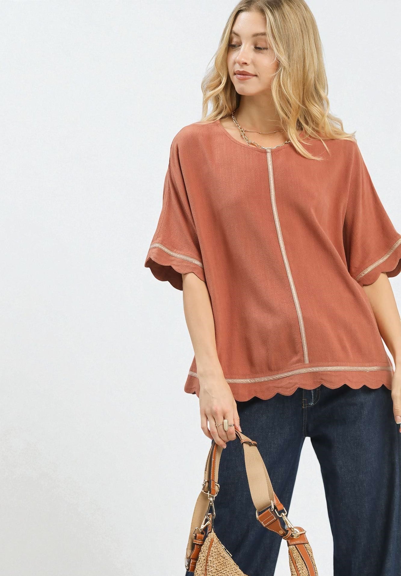 Woman wearing a rust-colored blouse with scalloped edges and dark jeans, holding a brown bag on a white background.