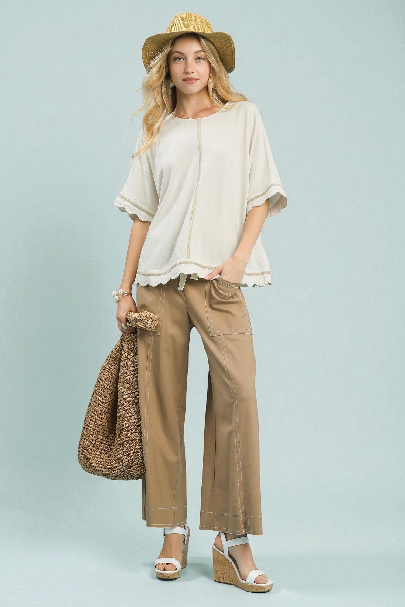 Woman wearing a beige top, tan pants, and a straw hat against a light blue background