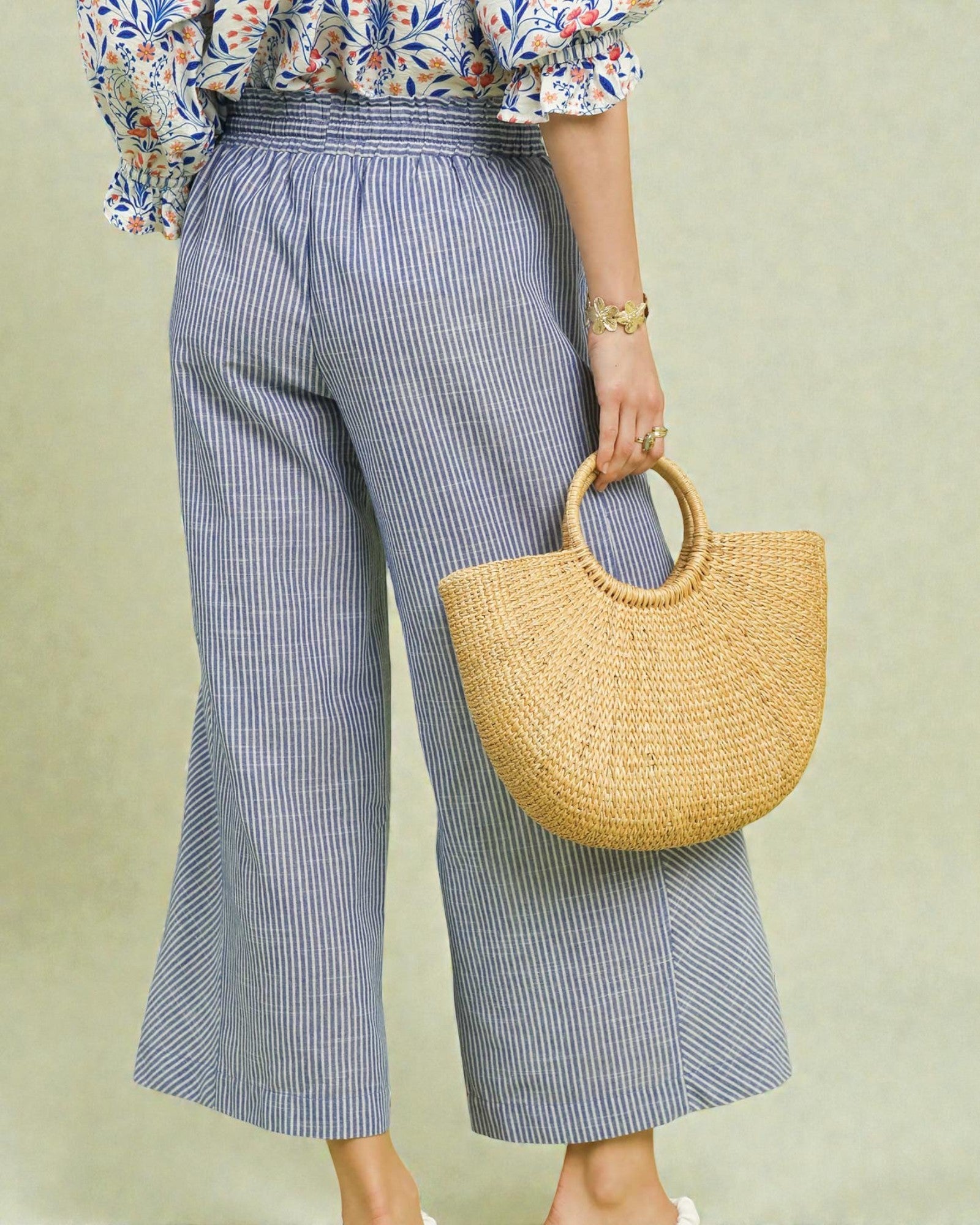 Person wearing blue striped pants and a floral top, holding a straw bag against a light green background