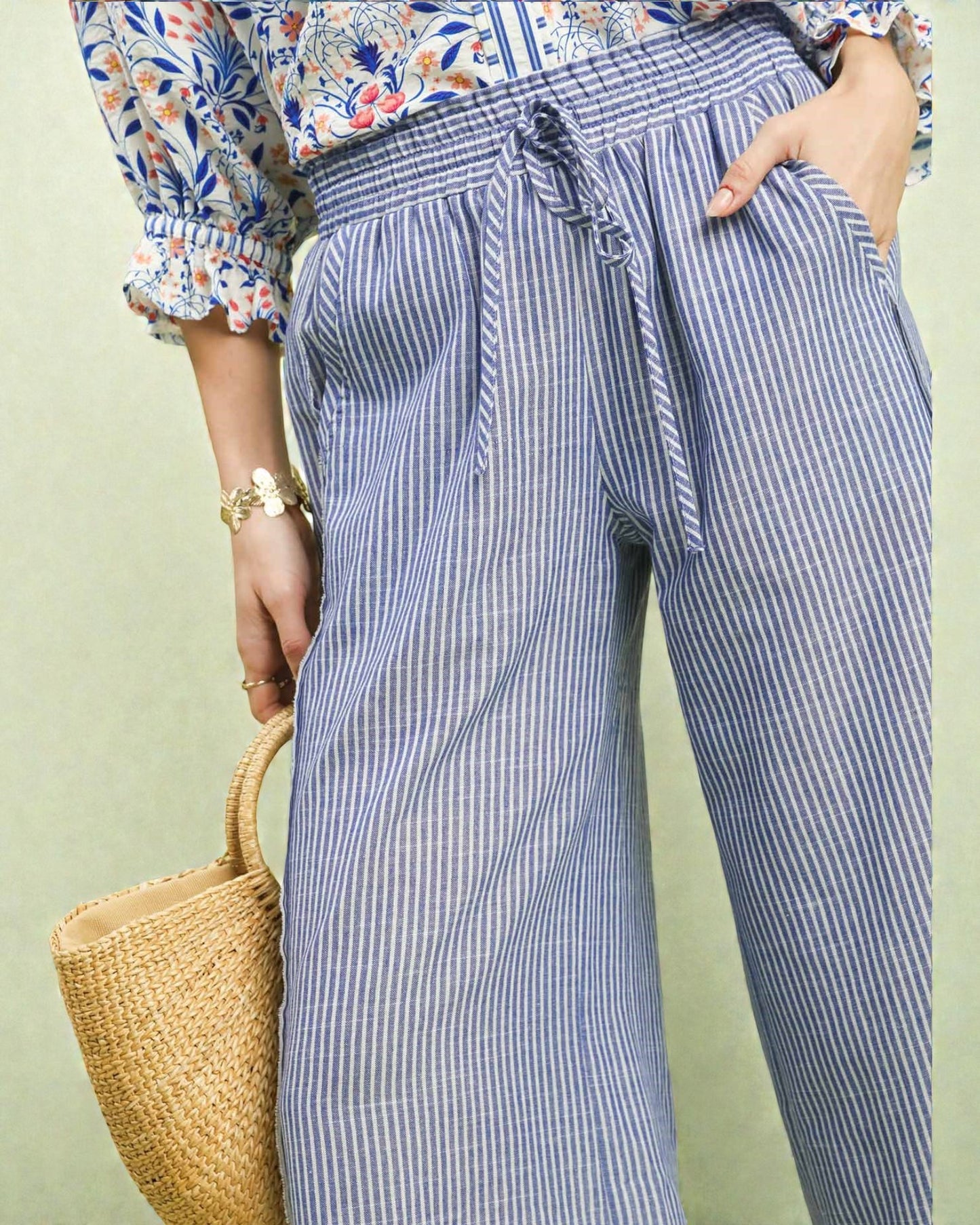 Person wearing blue striped pants with a floral blouse and holding a woven bag against a light green background