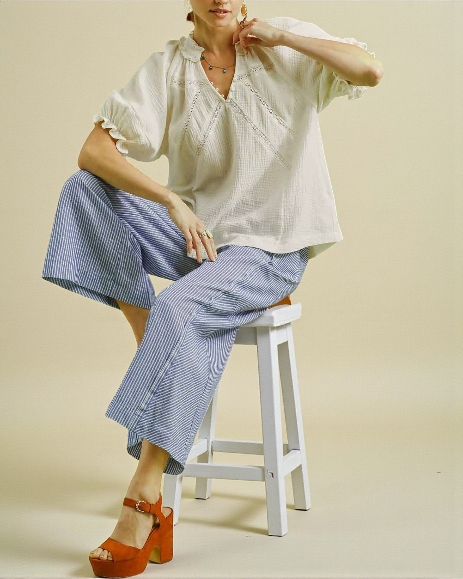 Woman sitting on a stool wearing a white blouse and blue striped pants with orange shoes.