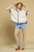 Woman wearing a white blouse with blue accents, denim shorts, and orange sandals against a beige background