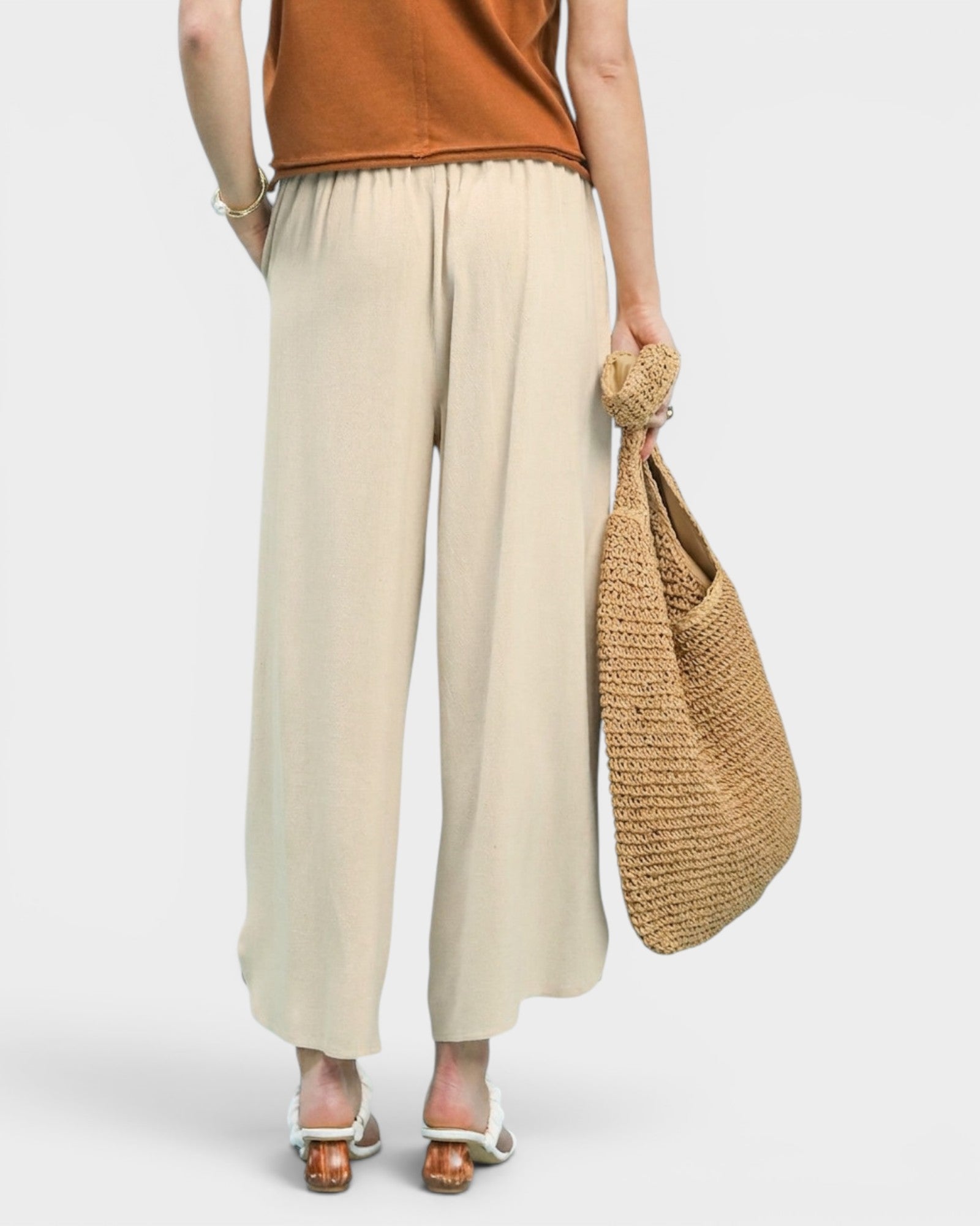 Person wearing beige wide-leg pants and a brown top, holding a woven bag on a white background