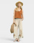 Woman wearing a rust-colored tank top and beige pants with floral embroidery, holding a straw bag on a white background.