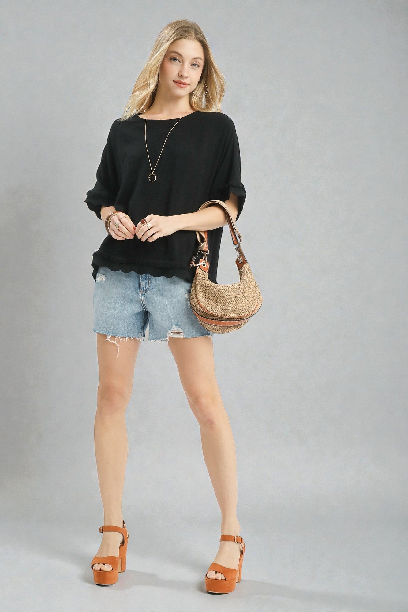 Woman wearing a black top, denim shorts, and brown sandals with a beige handbag on a gray background