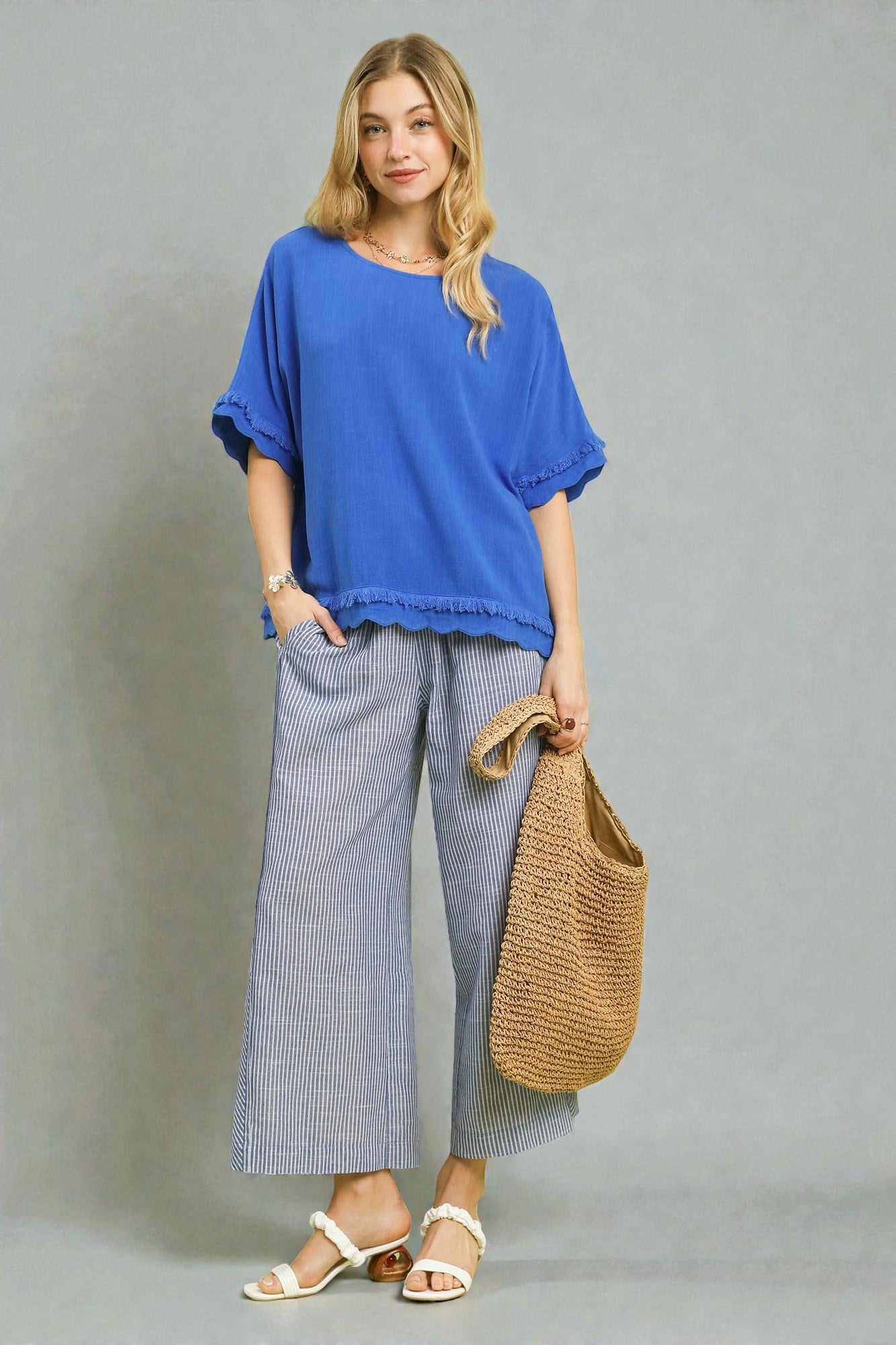 Woman wearing a blue top and gray pants, holding a woven bag against a gray background