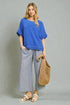 Woman wearing a blue top and gray pants, holding a woven bag against a gray background