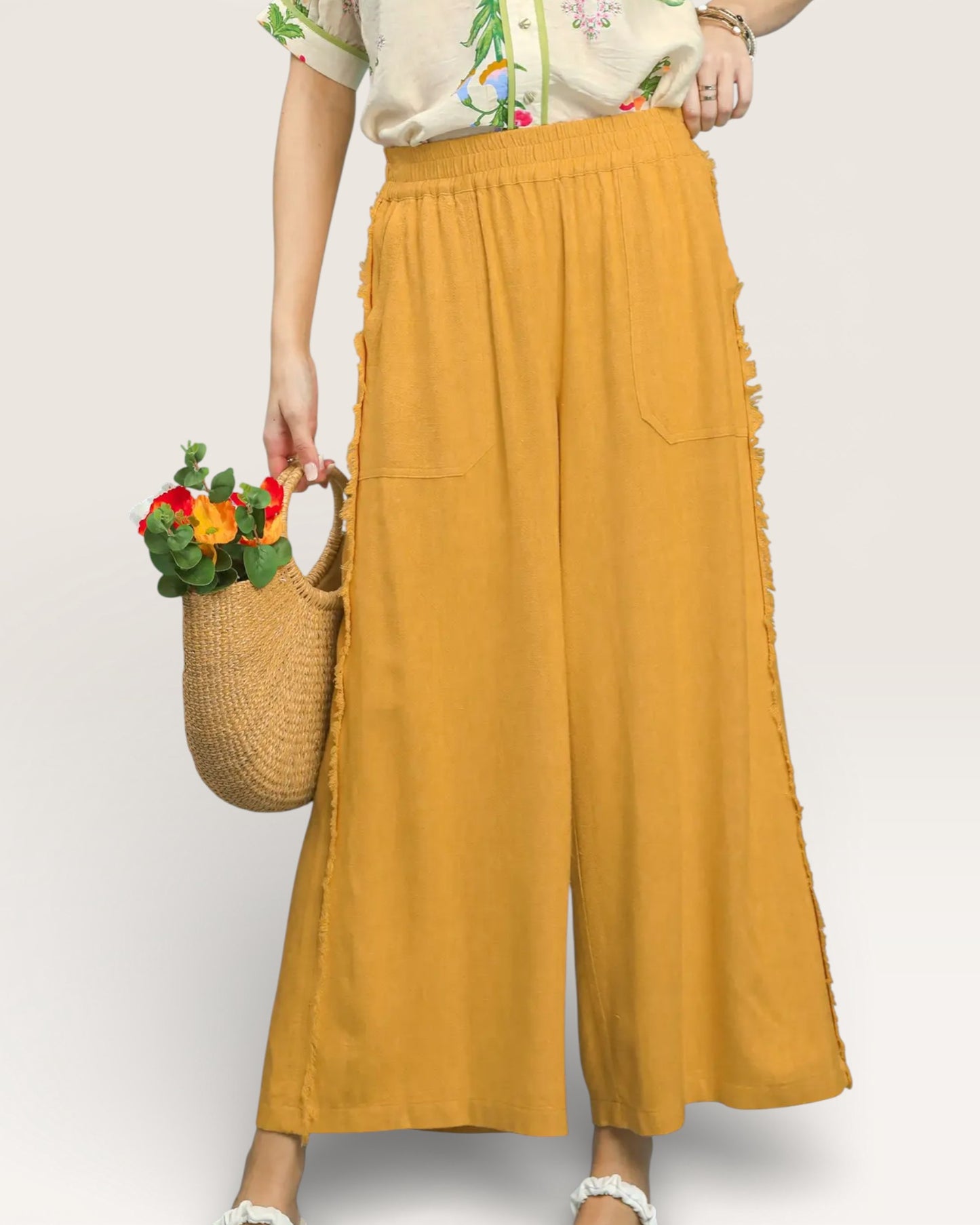 Person wearing mustard yellow wide-leg pants holding a woven bag with flowers on a white background