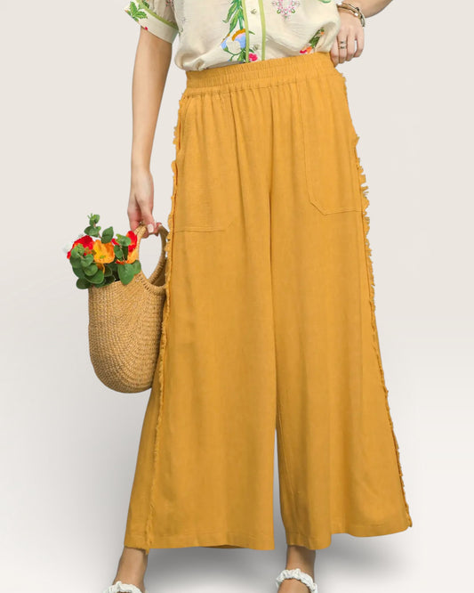 Person wearing mustard yellow wide-leg pants holding a woven bag with flowers on a white background