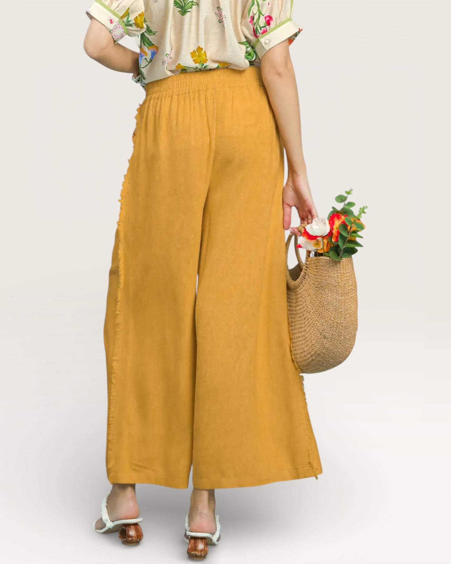 Person wearing mustard yellow wide-leg pants holding a woven bag with flowers on a plain background