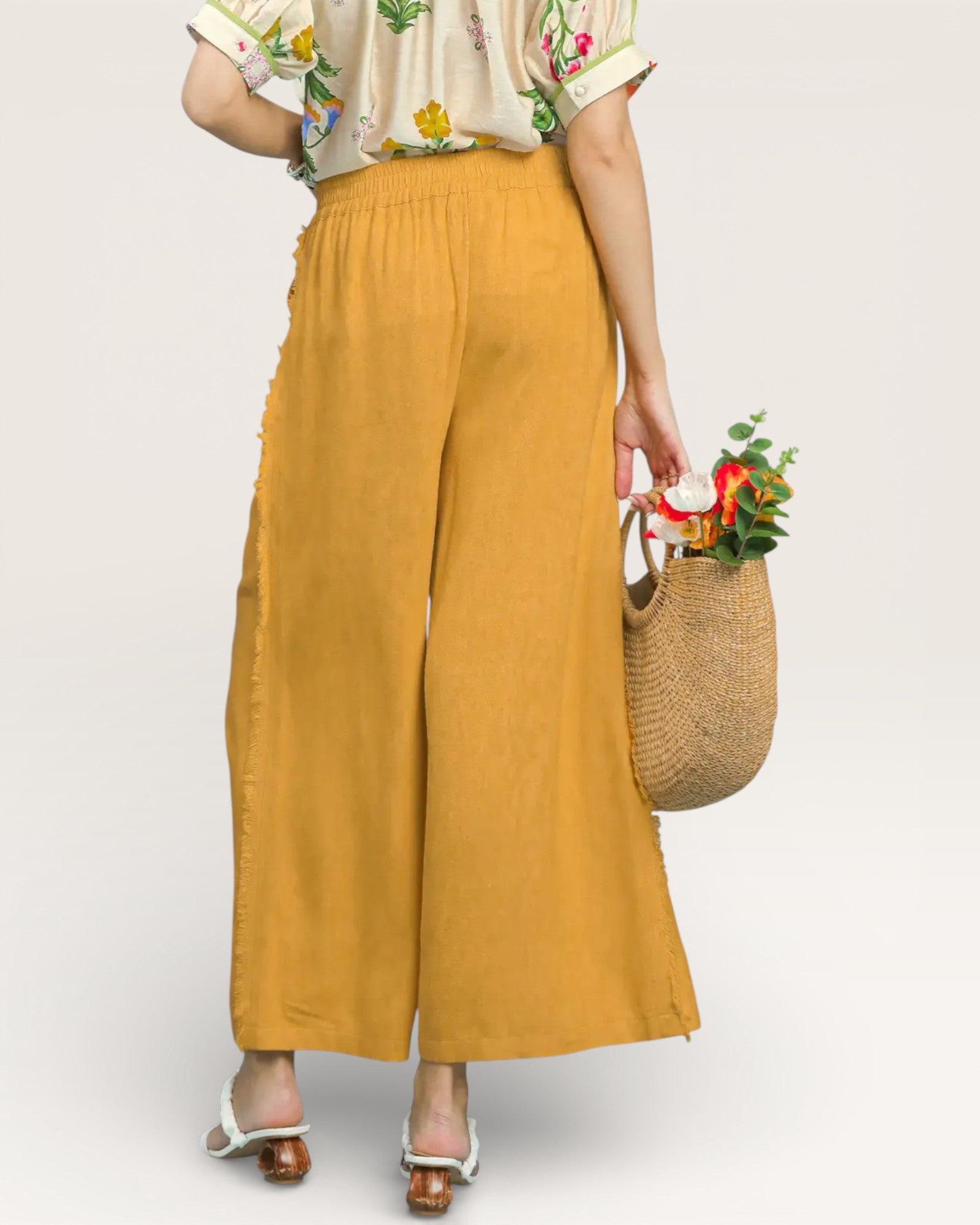 Person wearing mustard yellow wide-leg pants holding a woven bag with flowers on a plain background