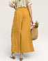 Person wearing mustard yellow wide-leg pants holding a woven bag with flowers on a plain background