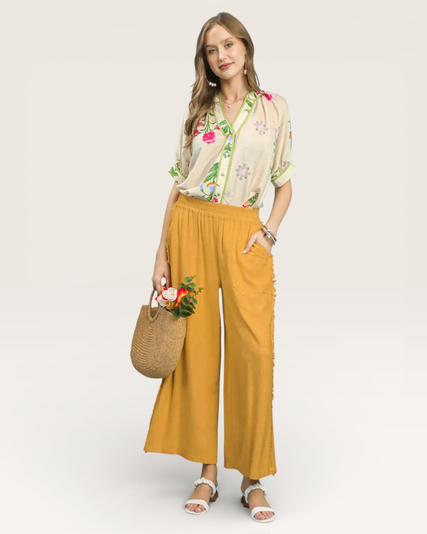 Woman wearing a floral blouse and mustard yellow pants with a straw bag on a white background