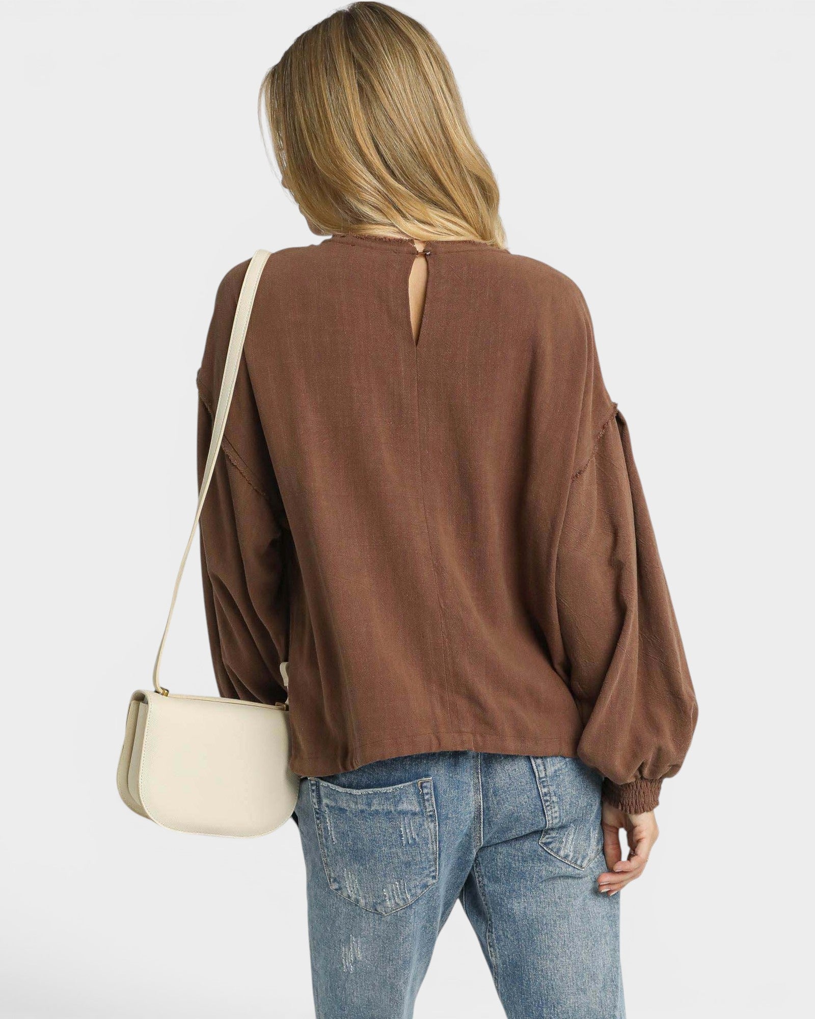 Person wearing a brown blouse with a cutout back design, holding a beige handbag on a white background.