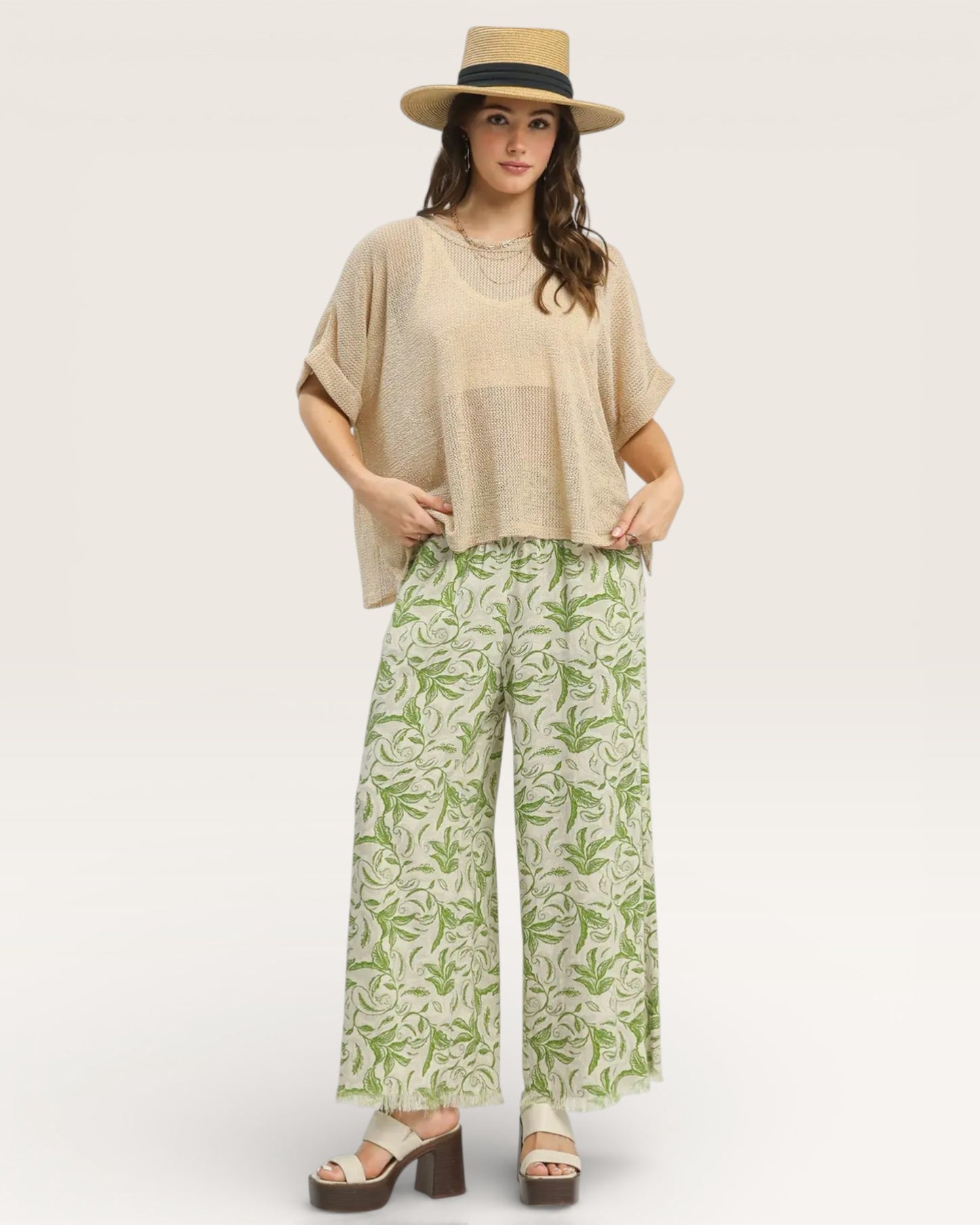 Woman wearing a beige top and green patterned pants with a hat on a white background