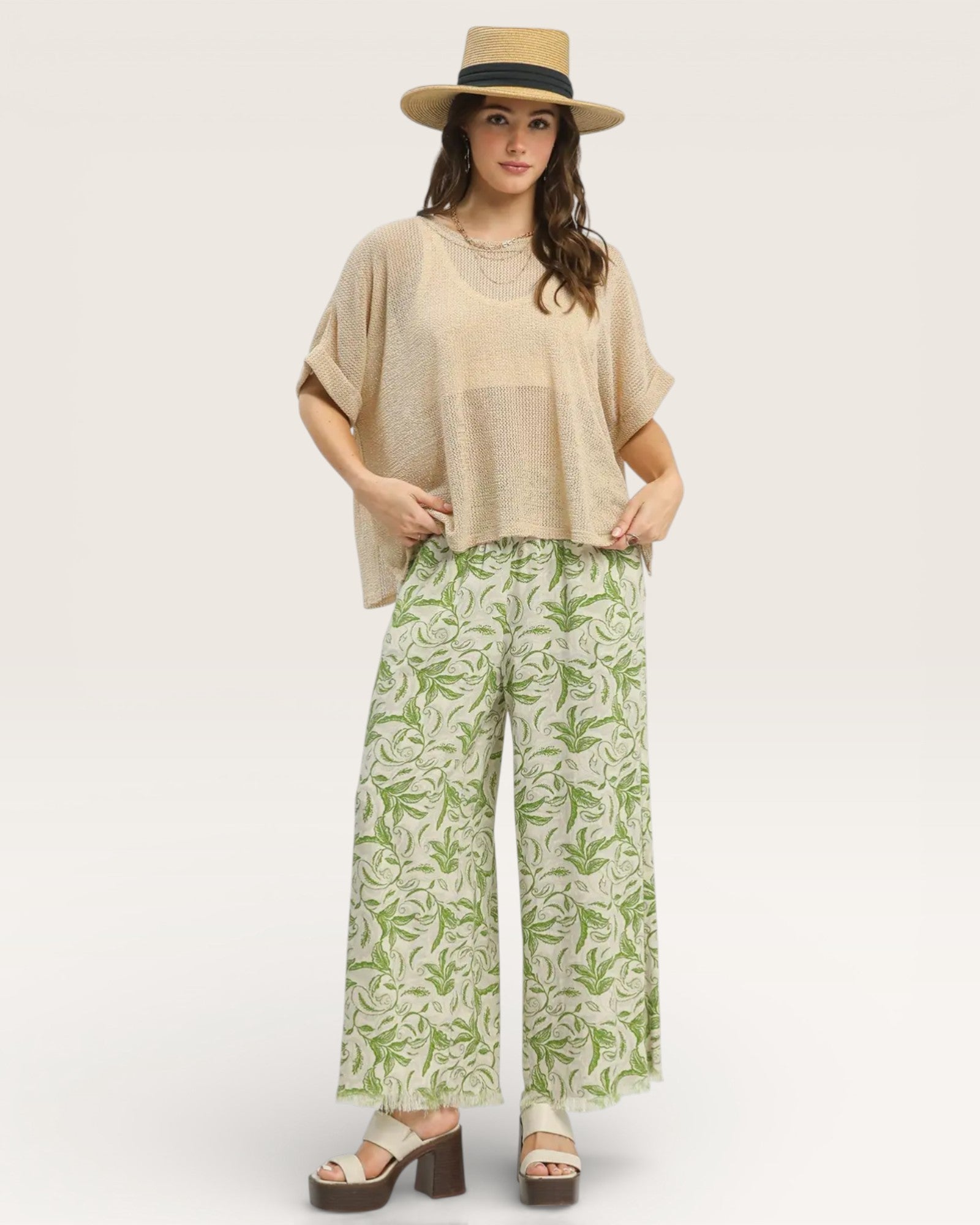 Woman wearing a beige top and green patterned pants with a hat on a white background