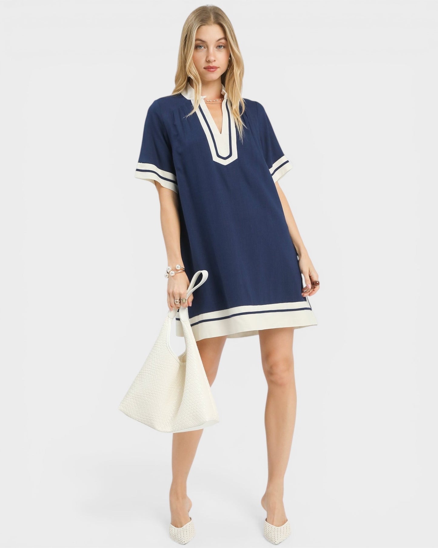 Woman wearing a navy dress with white trim holding a white handbag on a white background