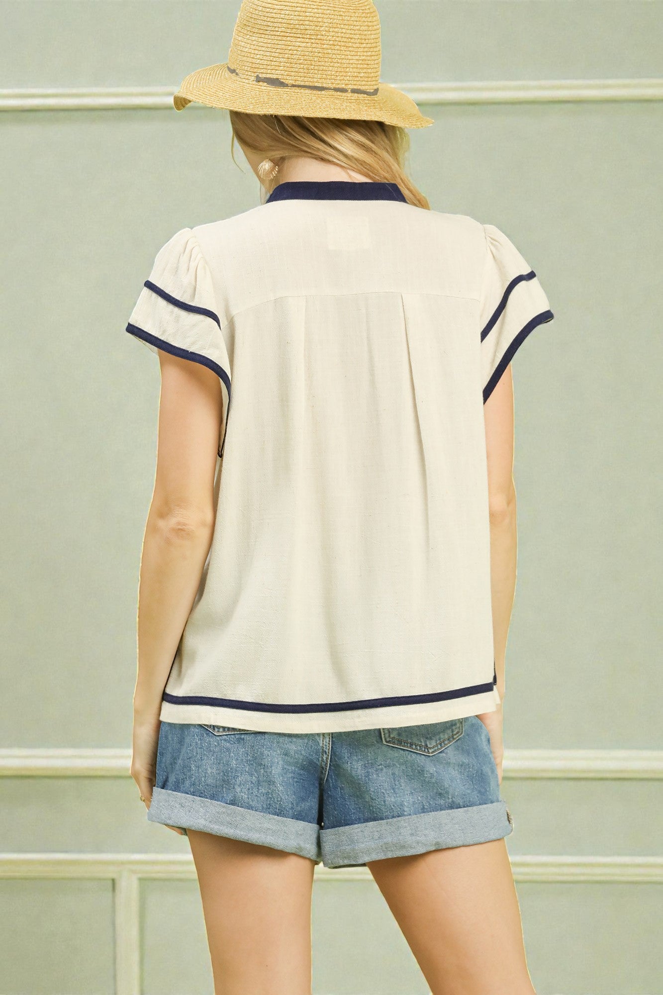 Person wearing a beige shirt with navy trim and denim shorts, standing against a light green wall.