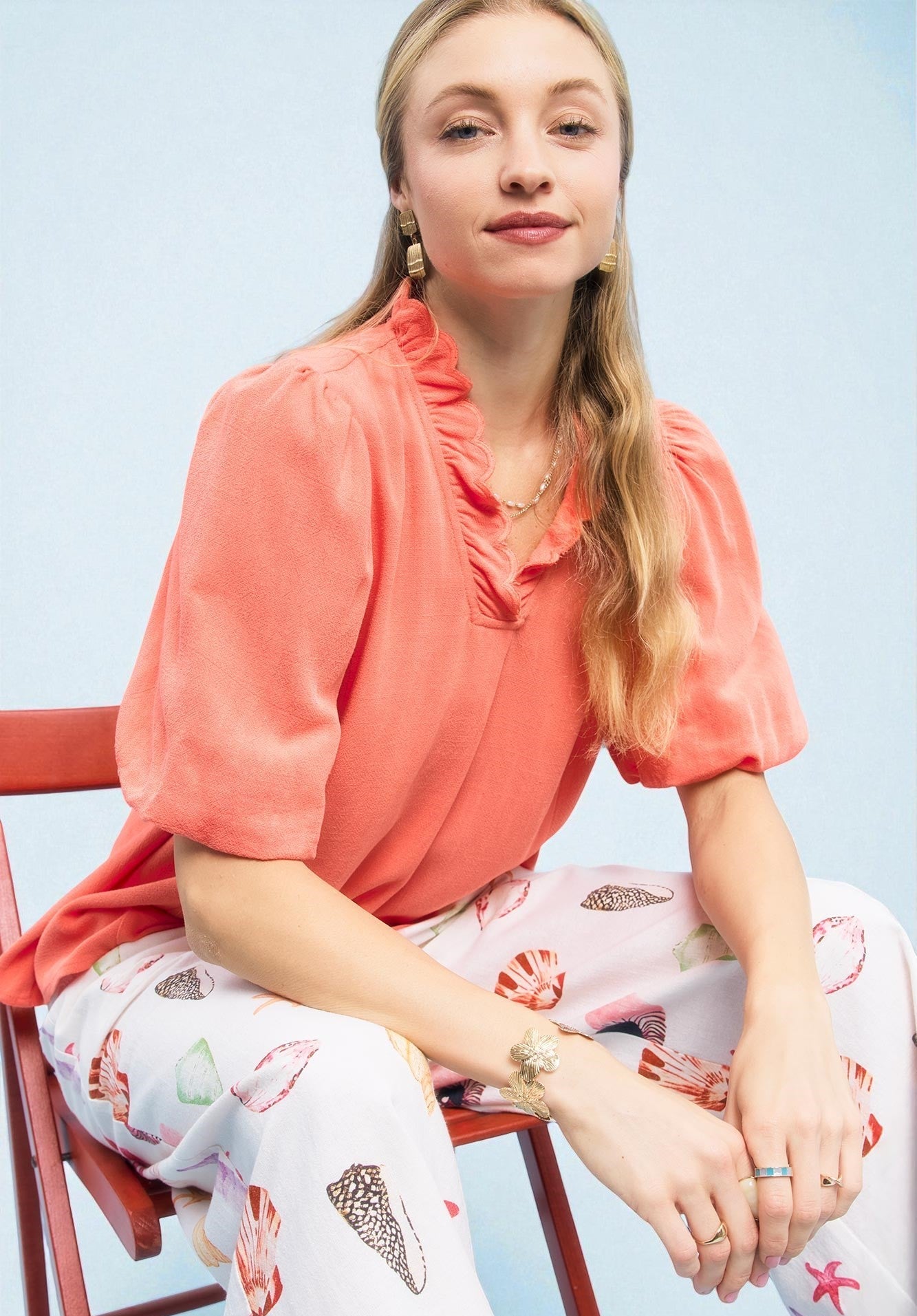 Woman wearing a coral top and floral pants sitting on a chair with a light blue background