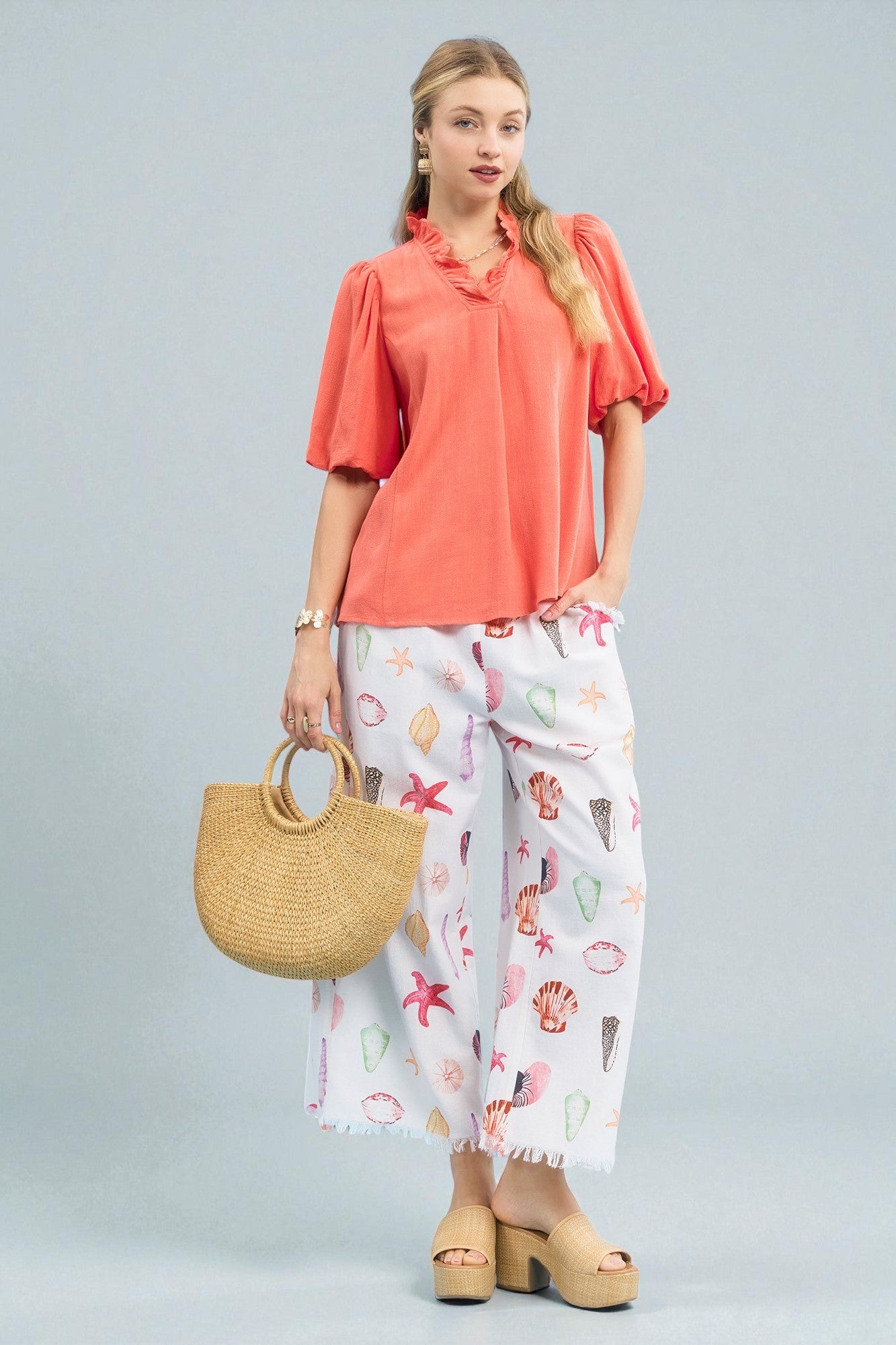 Woman wearing a coral blouse and white pants with shell pattern, holding a straw bag on a gray background