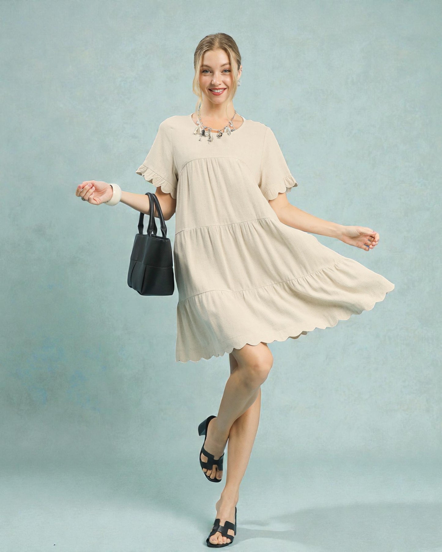 Woman in a beige dress holding a black handbag against a light blue background