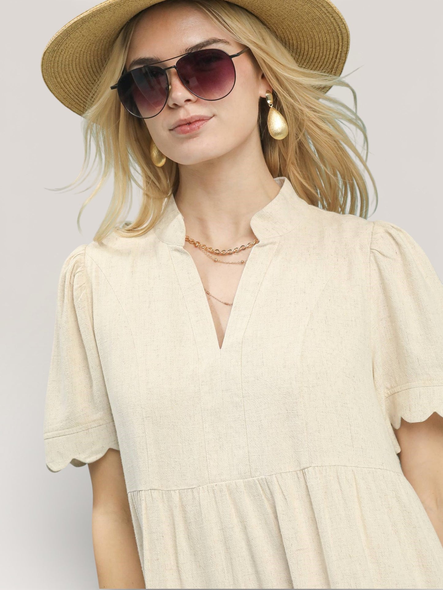 Woman wearing a beige dress, straw hat, and sunglasses on a plain background