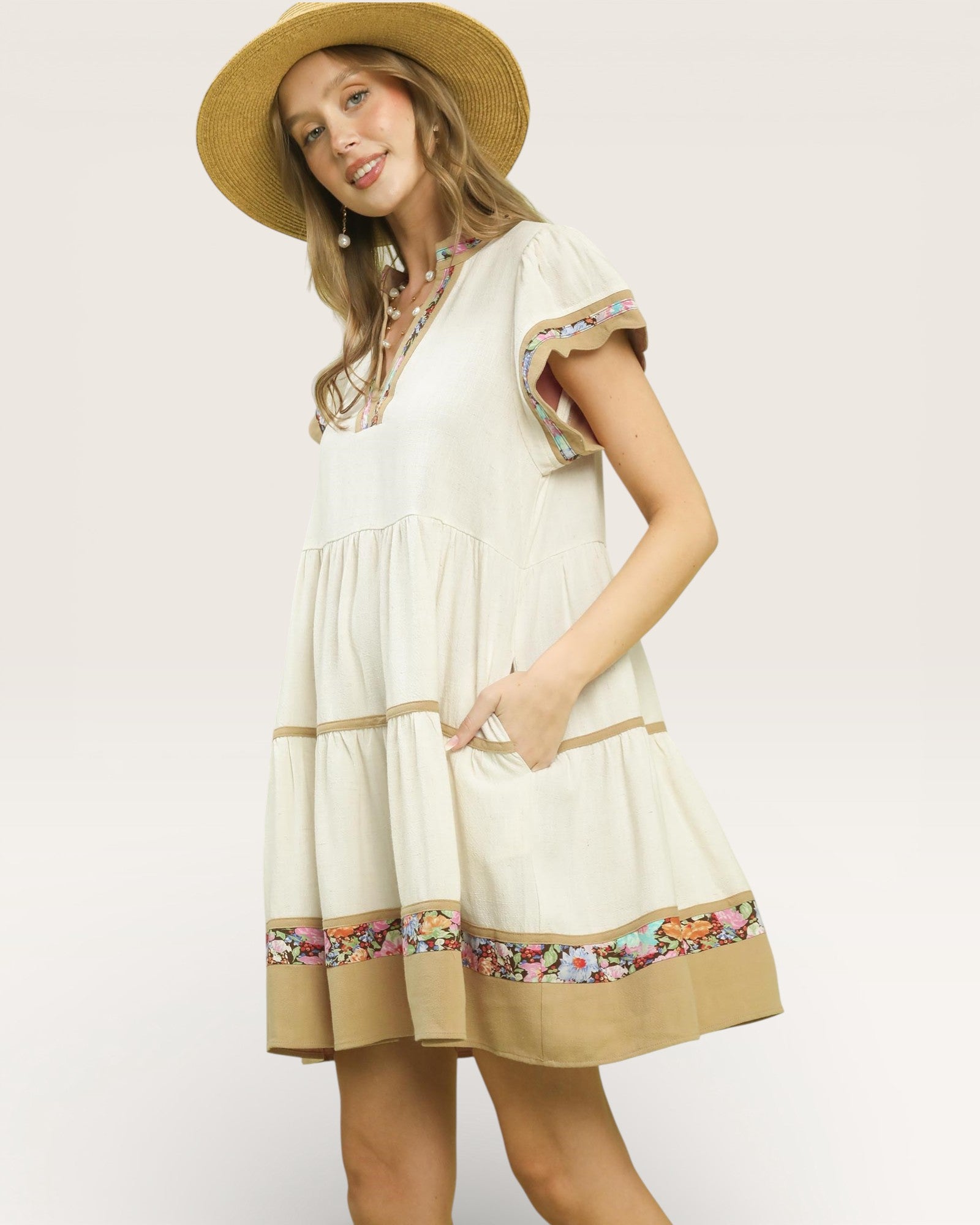 Woman wearing a white dress with floral trim and a straw hat on a plain background