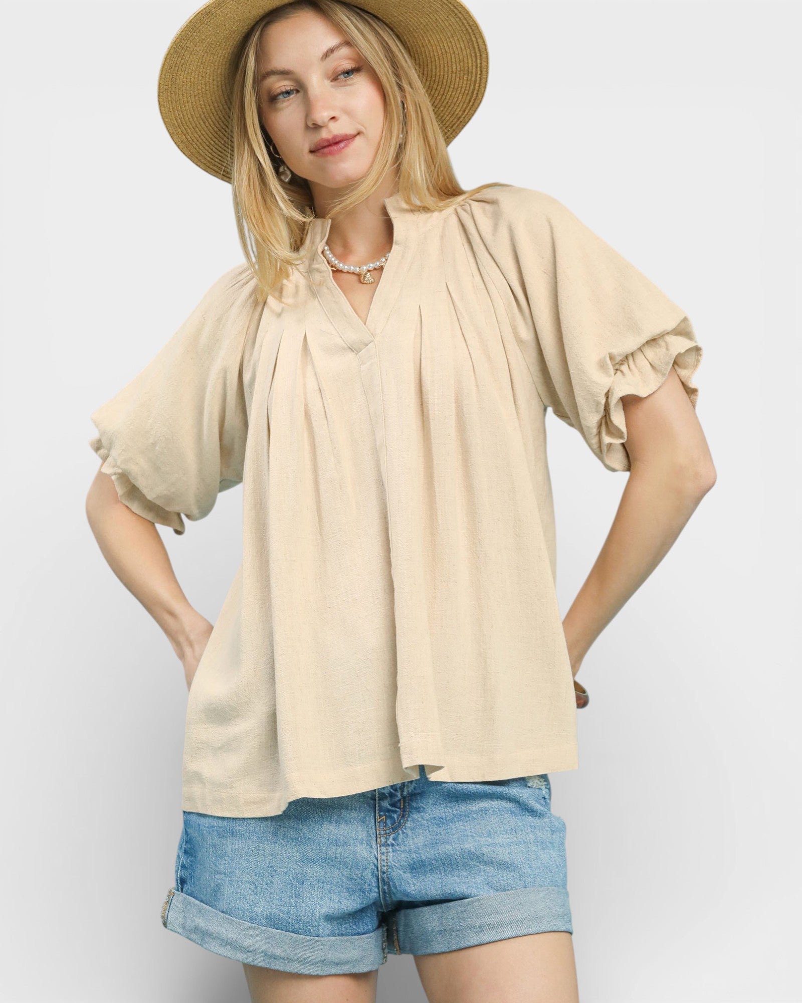 Woman wearing a beige blouse and denim shorts with a straw hat on a light background