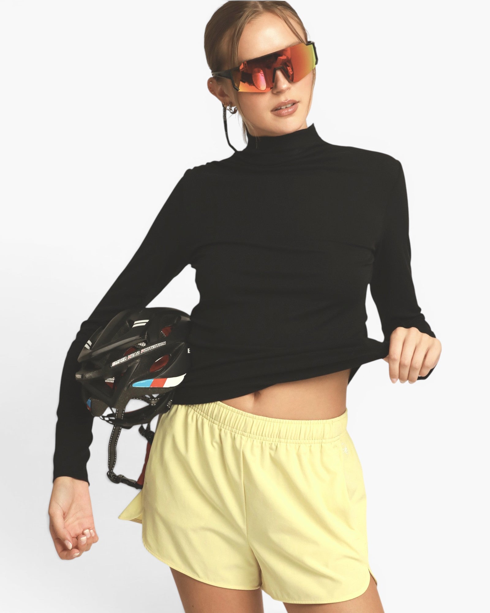 A woman modeling a black long-sleeve mock neck athletic top and light yellow shorts, holding a black bicycle helmet against a plain light gray studio background.