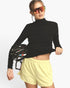 A woman modeling a black long-sleeve mock neck athletic top and light yellow shorts, holding a black bicycle helmet against a plain light gray studio background.