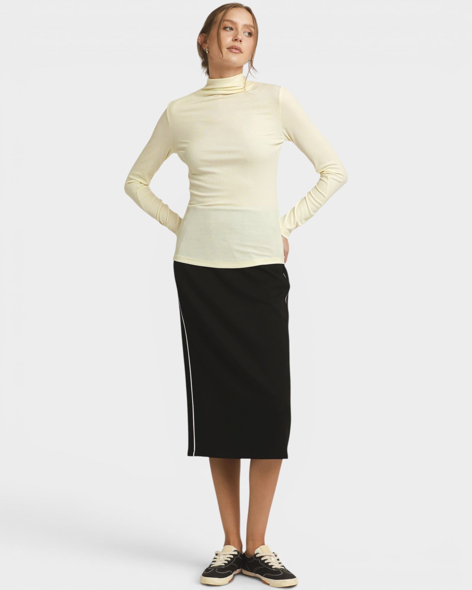 A woman modeling a cream ribbed mock neck long-sleeve top and a black midi skirt with white side stripes, paired with black and white sneakers, against a light gray studio background.