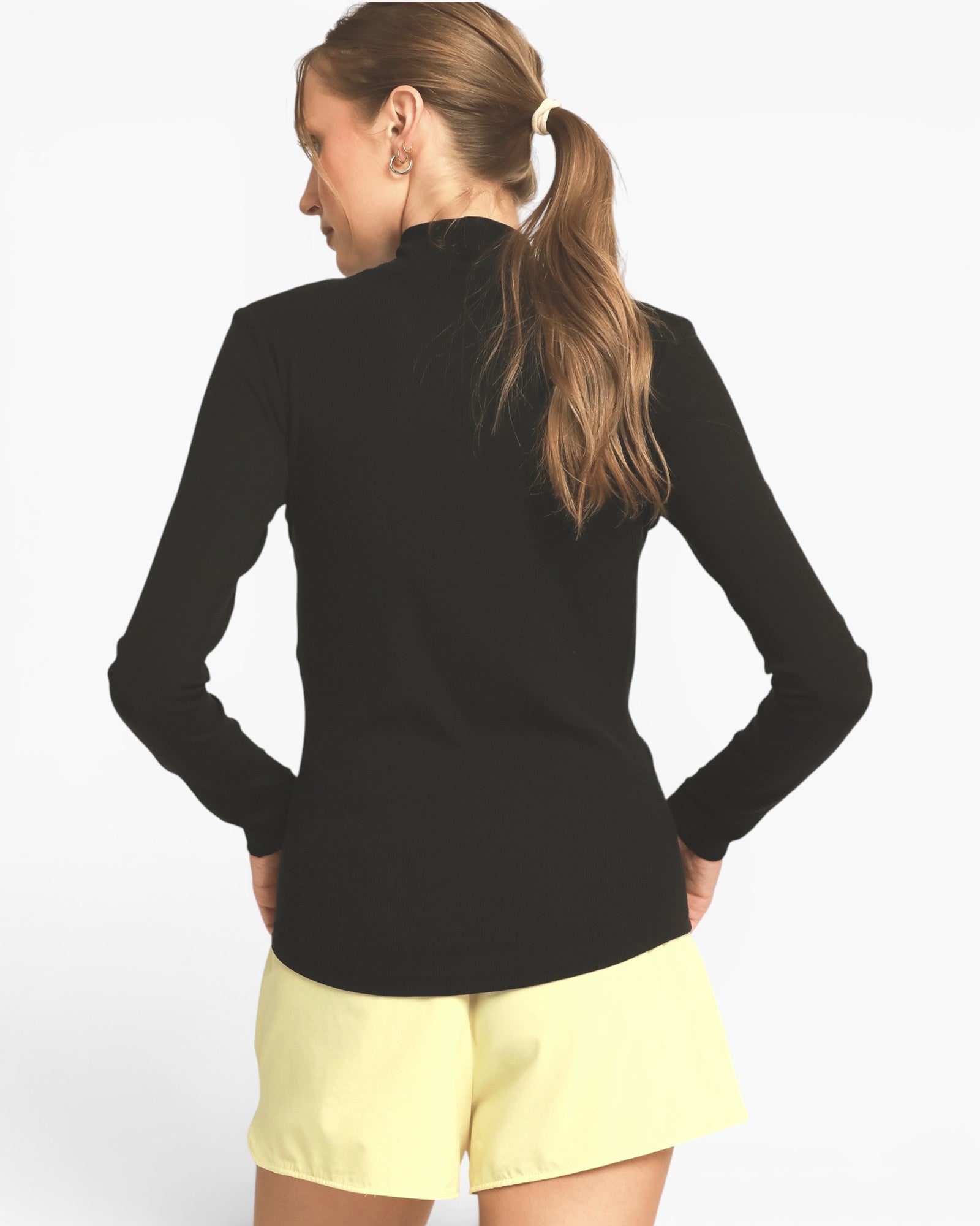 A woman shown from the back wearing a black long-sleeve mock neck top and light yellow shorts against a plain white background.