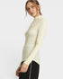 A woman wearing a fitted, long-sleeve mock neck top in a light cream color, paired with black pants, photographed against a solid light gray background.