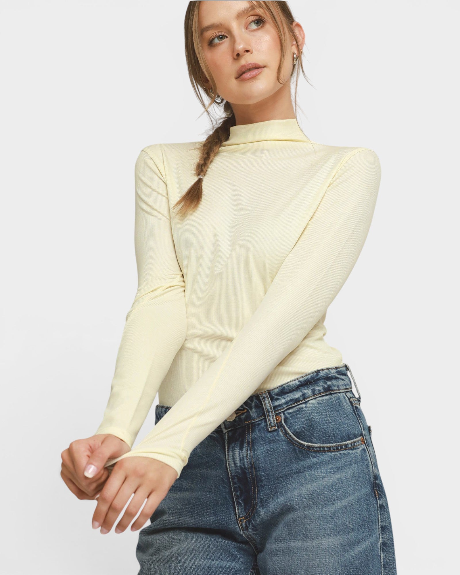 A woman wearing a pale yellow long-sleeve mock neck top and blue jeans, photographed against a light gray studio background.