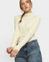 A woman wearing a pale yellow long-sleeve mock neck top and blue jeans, photographed against a light gray studio background.