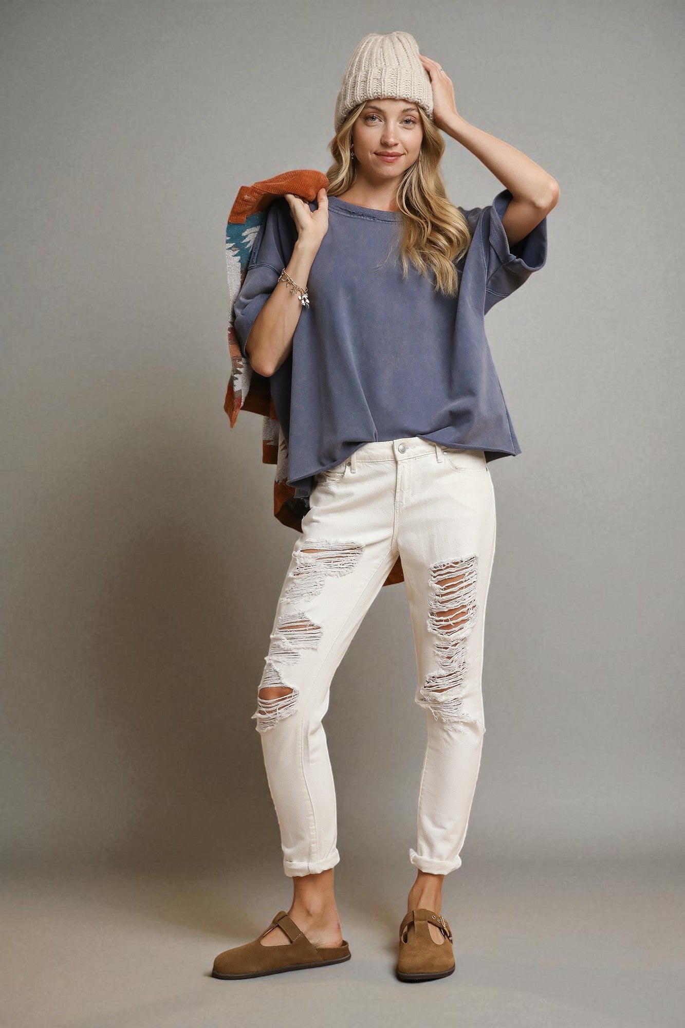 Woman wearing a blue shirt, white ripped jeans, and brown shoes against a gray background