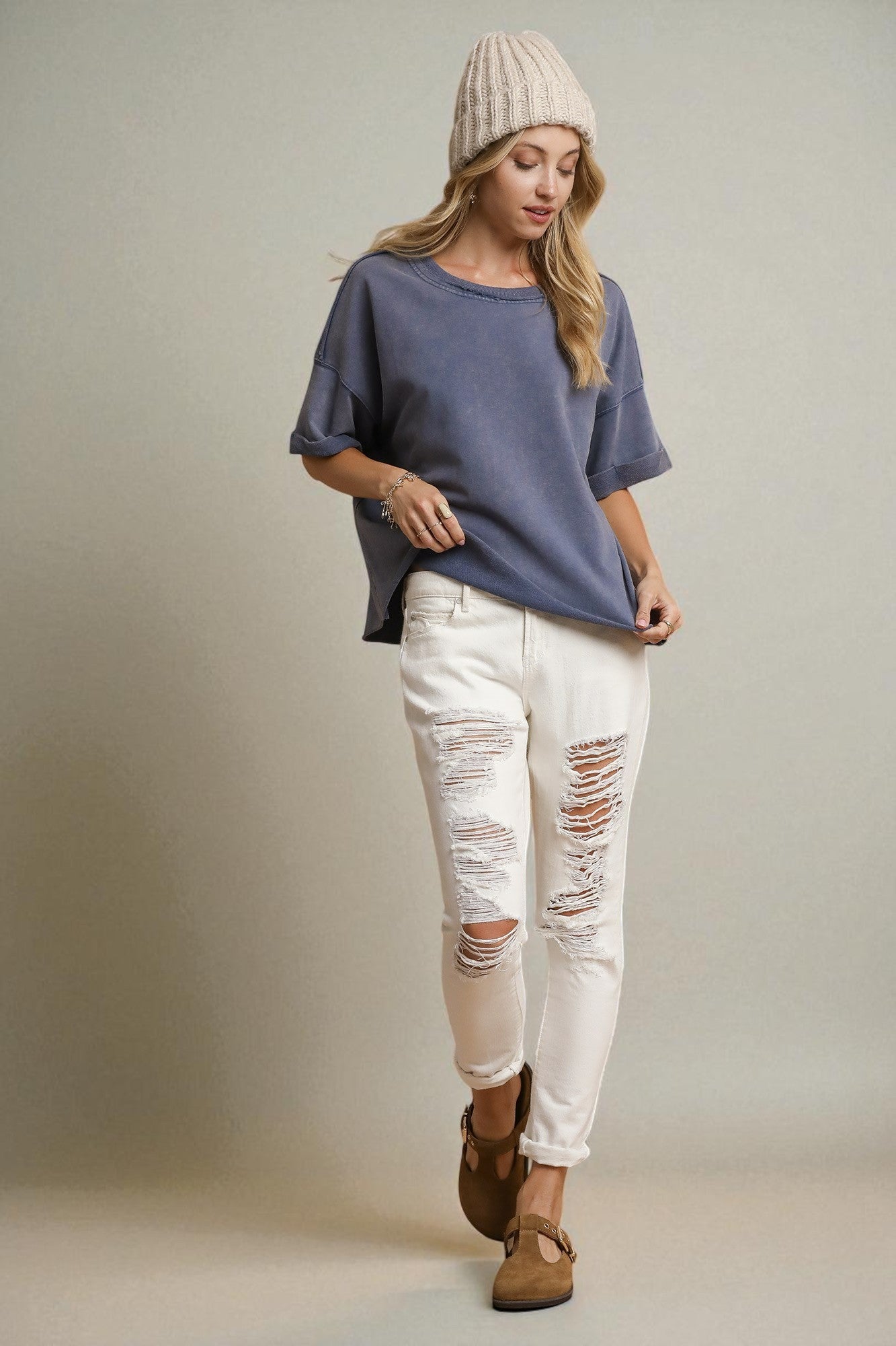 Woman wearing a blue shirt, white distressed jeans, and a beige beanie against a plain background