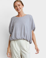 A woman wearing a light gray short-sleeve top with a textured waffle pattern and cream-colored pants, standing against a light gray studio background.