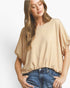 A woman wearing a beige short-sleeve top with a waffle texture and ruffled sleeves, paired with blue jeans, against a plain white background.