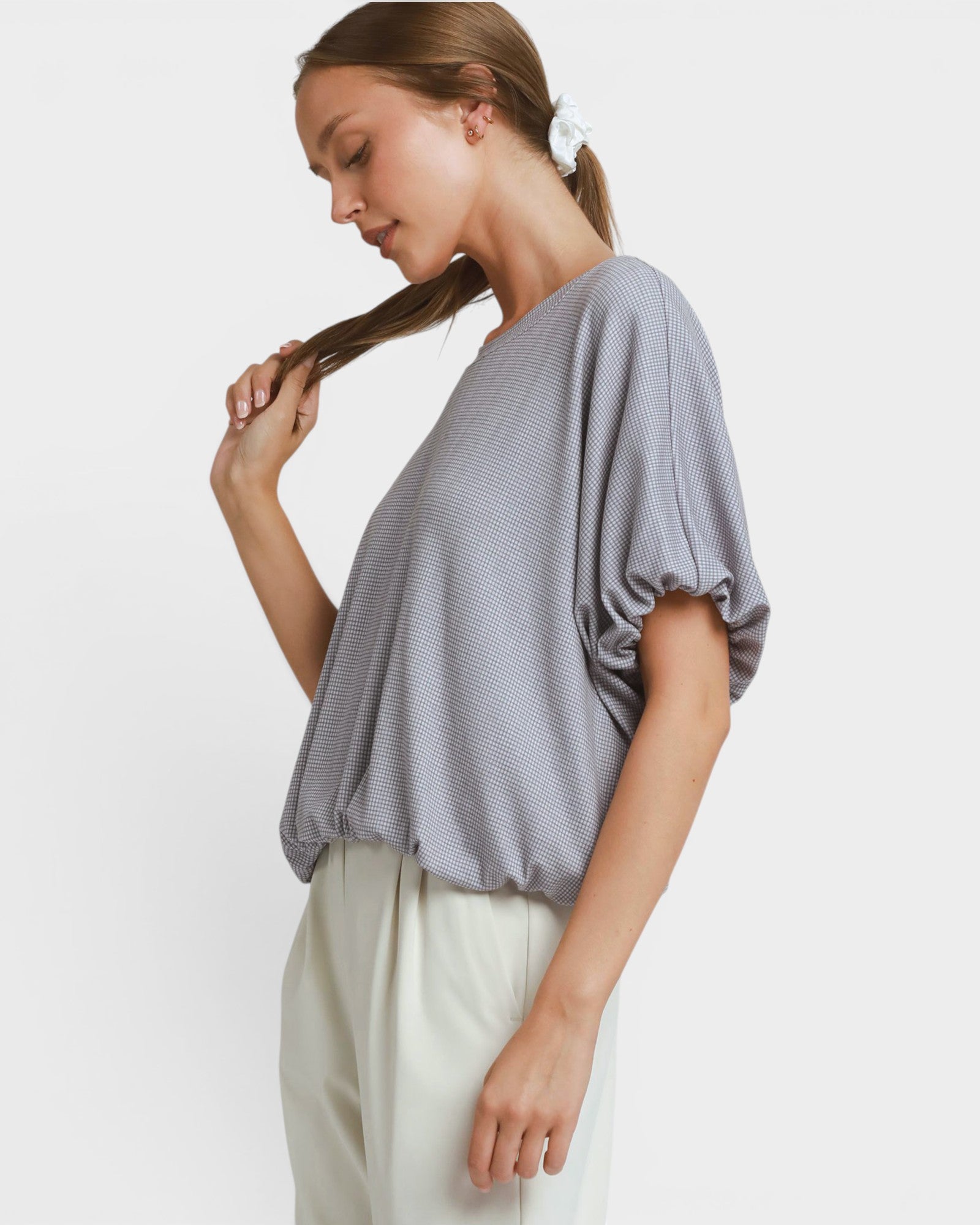 A woman wearing a light gray short-sleeve top with a textured pattern and cream-colored pants, shown from a side profile against a plain light gray background.