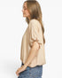 A woman shown in side profile wearing a light beige short-sleeve blouse with gathered puff sleeves and blue jeans, against a plain white studio background.