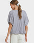 A woman shown from the back wearing a light gray and white micro-checkered short-sleeve top with a relaxed, balloon-style fit, paired with off-white pants against a plain light gray studio background.