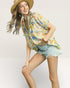 Woman wearing a floral shirt and denim shorts with a straw hat on a plain background