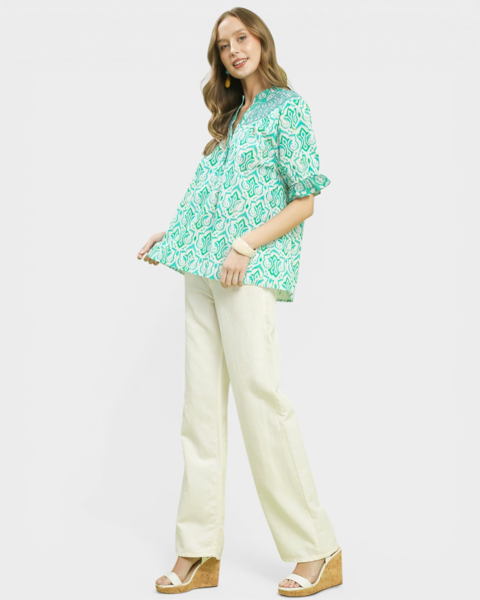 Woman wearing a green patterned top and white pants on a white background